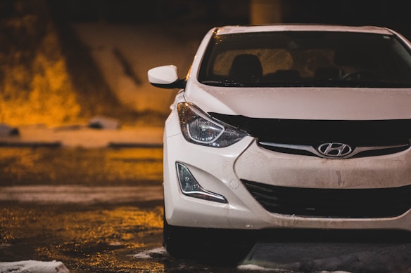 A white Hyundai car is parked in a dimly lit area with a wet surface from rainfall. The front of the car is prominently visible, showing the headlights and grille with the Hyundai logo. The background features blurred city lights, adding warmth to the scene.