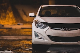 A white Hyundai car is parked in a dimly lit area with a wet surface from rainfall. The front of the car is prominently visible, showing the headlights and grille with the Hyundai logo. The background features blurred city lights, adding warmth to the scene.