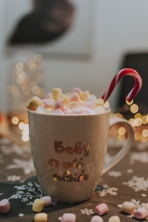A ceramic mug filled with small, multicolored marshmallows and a red and white candy cane. The mug is inscribed with the phrase 'Baby it's cold outside' and is set on a dark cloth with white snowflake patterns. In the background, soft, warm lights create a cozy, festive atmosphere.