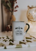 A white mug with the phrase 'Merry Christmas' printed in festive typography, accompanied by small images of reindeer and a tree. The mug is placed on a cloth with green and brown tree patterns. The background is softly lit, with hints of holiday decor such as pine branches and berries.
