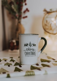 A white mug with the phrase 'Merry Christmas' printed in festive typography, accompanied by small images of reindeer and a tree. The mug is placed on a cloth with green and brown tree patterns. The background is softly lit, with hints of holiday decor such as pine branches and berries.