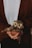 A person is holding a small, handcrafted owl made from natural materials such as pine cones and straw. The owl is adorned with a colorful woven scarf. The person is wearing a brown knitted cardigan over a white shirt.