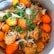 A pot filled with a vibrant stew comprising chicken pieces, whole orange tomatoes, and topped with fresh garnish like cilantro and chopped green onions. The ingredients are cooked in a rich broth that seems to enhance the color of the vegetables and meat.