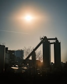 A silhouette of an industrial facility is set against a bright sun in the sky. The scene includes tall structures with a conveyor belt and some storage tanks around them. The sunlight casts long shadows, and a bare tree is visible in the foreground.