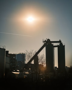 A silhouette of an industrial facility is set against a bright sun in the sky. The scene includes tall structures with a conveyor belt and some storage tanks around them. The sunlight casts long shadows, and a bare tree is visible in the foreground.