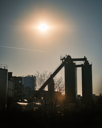 A silhouette of an industrial facility is set against a bright sun in the sky. The scene includes tall structures with a conveyor belt and some storage tanks around them. The sunlight casts long shadows, and a bare tree is visible in the foreground.