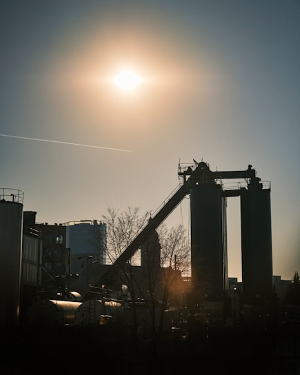 A silhouette of an industrial facility is set against a bright sun in the sky. The scene includes tall structures with a conveyor belt and some storage tanks around them. The sunlight casts long shadows, and a bare tree is visible in the foreground.
