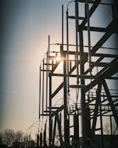 A silhouetted structure of metallic electrical components set against the backdrop of a setting or rising sun. The structure features tall vertical rods and various connected insulators, with the sun creating a bright glare through the framework. Bare trees are visible in the foreground, adding to the industrial setting.