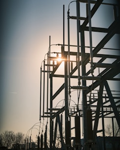 A silhouetted structure of metallic electrical components set against the backdrop of a setting or rising sun. The structure features tall vertical rods and various connected insulators, with the sun creating a bright glare through the framework. Bare trees are visible in the foreground, adding to the industrial setting.