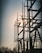 A silhouetted structure of metallic electrical components set against the backdrop of a setting or rising sun. The structure features tall vertical rods and various connected insulators, with the sun creating a bright glare through the framework. Bare trees are visible in the foreground, adding to the industrial setting.
