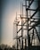 A silhouetted structure of metallic electrical components set against the backdrop of a setting or rising sun. The structure features tall vertical rods and various connected insulators, with the sun creating a bright glare through the framework. Bare trees are visible in the foreground, adding to the industrial setting.