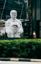 A large, intricately designed sculpture composed of white letters arranged to form a human figure is prominently displayed in a modern urban setting. The sculpture sits on a pedestal surrounded by lush green shrubs, with a building featuring glass windows visible in the background. A person walking nearby adds scale to the scene.
