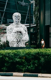 A large, intricately designed sculpture composed of white letters arranged to form a human figure is prominently displayed in a modern urban setting. The sculpture sits on a pedestal surrounded by lush green shrubs, with a building featuring glass windows visible in the background. A person walking nearby adds scale to the scene.
