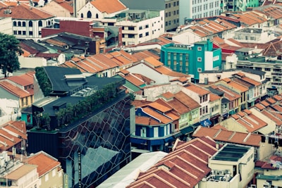 A vibrant cityscape featuring a mix of modern and traditional architecture. Numerous buildings with red-tiled roofs fill the scene, complemented by a striking modern building with a glass exterior and rooftop greenery. The juxtaposition of old and new structures creates a dynamic urban environment.