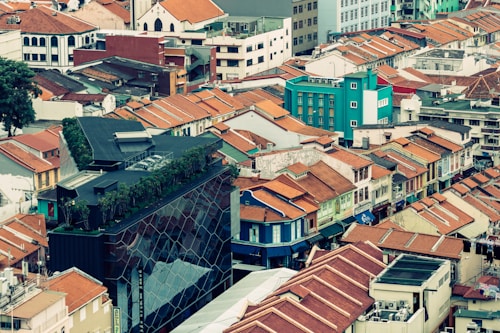 A vibrant cityscape featuring a mix of modern and traditional architecture. Numerous buildings with red-tiled roofs fill the scene, complemented by a striking modern building with a glass exterior and rooftop greenery. The juxtaposition of old and new structures creates a dynamic urban environment.