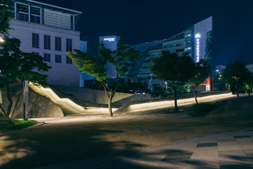 An urban setting at night features modern architecture with illuminated pathways and sleek building designs. Trees and landscaped areas are interspersed throughout the scene, providing a serene contrast to the contemporary structures. Soft lighting accentuates the geometric lines and architectural details.