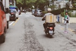 In an urban street setting, a person rides a scooter carrying a large cardboard box on the back. Vehicles line the road on either side while a pedestrian stands on the right sidewalk, dressed casually and holding a phone. The scene is populated with buildings, trees, and parked cars, reflecting a typical city environment.
