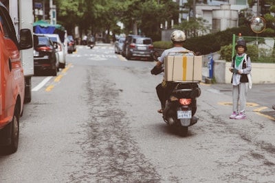 In an urban street setting, a person rides a scooter carrying a large cardboard box on the back. Vehicles line the road on either side while a pedestrian stands on the right sidewalk, dressed casually and holding a phone. The scene is populated with buildings, trees, and parked cars, reflecting a typical city environment.