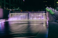A nighttime urban waterfall installation with vibrant lighting illuminating the water in shades of purple and white. It is situated next to a set of concrete stairs with metal railings, and there is greenery beside the staircase. Tall buildings are partially visible in the background, contributing to a cityscape atmosphere.
