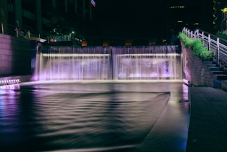 A nighttime urban waterfall installation with vibrant lighting illuminating the water in shades of purple and white. It is situated next to a set of concrete stairs with metal railings, and there is greenery beside the staircase. Tall buildings are partially visible in the background, contributing to a cityscape atmosphere.