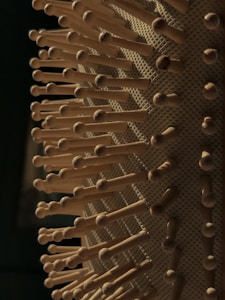 Close-up of a hairbrush with wooden bristles, each evenly spaced and protruding from a textured base. The lighting casts soft shadows, highlighting the natural wood tones and pattern of the brush.