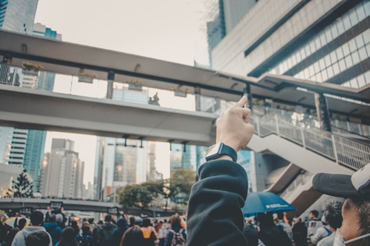 A crowd of people gathered in an urban environment with tall buildings in the background. A raised hand, wearing a smartwatch, is prominently visible in the foreground, suggesting a demonstration or protest. An overpass with people standing on it extends across the image.