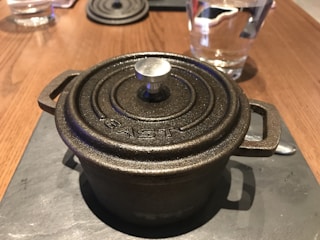 A cast iron pot with a circular lid rests on a dark surface, with a knob in the center and the word 'EAST' embossed on the top. In the background, there is a glass of water and a spoon on a wooden table.