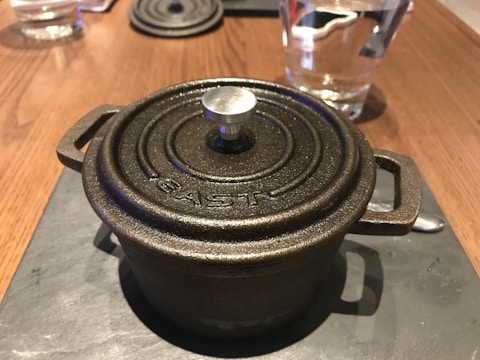 A cast iron pot with a circular lid rests on a dark surface, with a knob in the center and the word 'EAST' embossed on the top. In the background, there is a glass of water and a spoon on a wooden table.