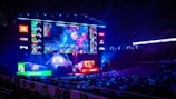 A large esports arena featuring a massive screen displaying a video game, surrounded by sponsor logos and team names. The auditorium is filled with spectators watching the gaming event. Neon blue and purple lights illuminate the stage, creating a vibrant atmosphere.