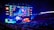 A large esports arena featuring a massive screen displaying a video game, surrounded by sponsor logos and team names. The auditorium is filled with spectators watching the gaming event. Neon blue and purple lights illuminate the stage, creating a vibrant atmosphere.