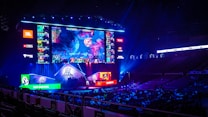 A large esports arena featuring a massive screen displaying a video game, surrounded by sponsor logos and team names. The auditorium is filled with spectators watching the gaming event. Neon blue and purple lights illuminate the stage, creating a vibrant atmosphere.