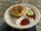 A white disposable plate holds a sesame seed hamburger bun representing a sandwich, accompanied by sauces in separate compartments. The sauces include a red ketchup, a green mint chutney, and a brown chutney. The background features a blurred, textured stone surface, and the plate is resting on a person's knee.