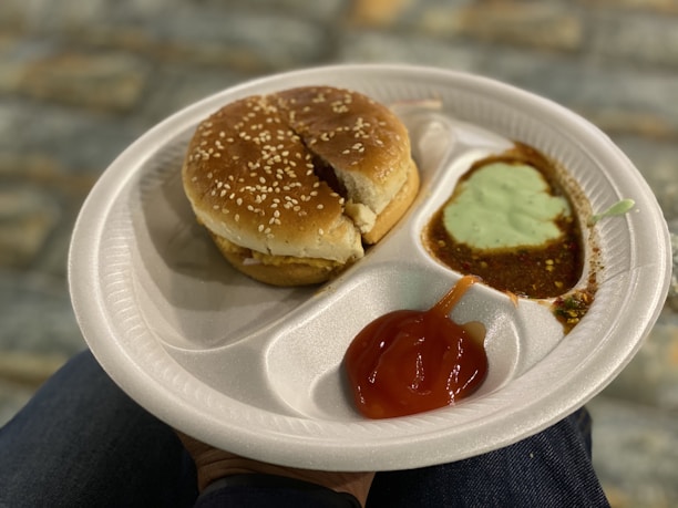 A white disposable plate holds a sesame seed hamburger bun representing a sandwich, accompanied by sauces in separate compartments. The sauces include a red ketchup, a green mint chutney, and a brown chutney. The background features a blurred, textured stone surface, and the plate is resting on a person's knee.