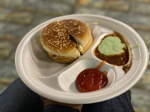 A white disposable plate holds a sesame seed hamburger bun representing a sandwich, accompanied by sauces in separate compartments. The sauces include a red ketchup, a green mint chutney, and a brown chutney. The background features a blurred, textured stone surface, and the plate is resting on a person's knee.