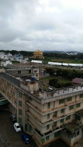 An urban landscape with a large multi-story building in the foreground. The building has a flat roof and several rows of windows with a neutral color scheme. Surrounding the building are other structures, including a recognizable golden temple with intricate designs amidst greenery and hills in the background. The sky is overcast, creating a muted atmosphere.