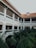 A two-story building with white walls, green railings, and red-tiled roofs surrounds a courtyard filled with lush green palm trees. The upper level features a series of archways and columns, and windows are equipped with beige shutters.
