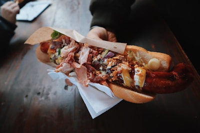 A person holds a large, loaded hot dog topped with various ingredients such as bacon, fried onions, mayonnaise, mustard, pickles, and other condiments. The hot dog is wrapped in paper and held over a napkin, with a wooden table in the background.