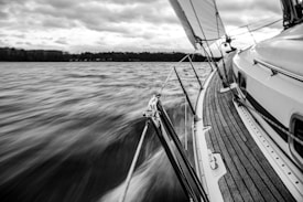 A monochromatic image capturing the dynamic motion of a sailboat cutting through water. The deck of the boat, made of smooth wood, is shot from a low angle as it leans with the wind. The sails are full, and the blurred water flows past, indicating speed. A dramatic sky with heavy clouds looms overhead.