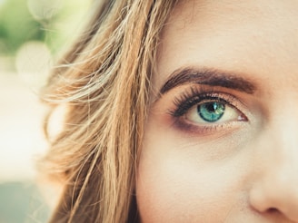 A close-up of a person's face with a focus on the right eye, showcasing vibrant blue iris and detailed eyelashes. The surrounding skin appears smooth with natural makeup, and strands of light brown hair frame the face.