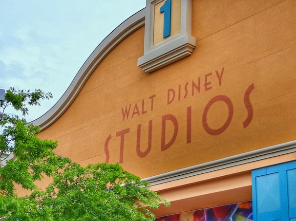 A section of a building showcasing a large sign that reads 'Walt Disney Studios'. The architectural design features a curved, decorative top and a number '1' displayed in a blue square. To the left, a tree branch with green leaves contrasts against the building's warm tan color.