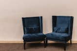 A pair of plush, dark blue velvet armchairs with high backs are positioned on a carpeted floor against a plain light beige wall. The chairs have decorative studded trim along the edges and are accompanied by matching lumbar pillows placed on the seat.