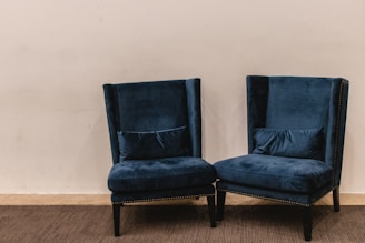 A pair of plush, dark blue velvet armchairs with high backs are positioned on a carpeted floor against a plain light beige wall. The chairs have decorative studded trim along the edges and are accompanied by matching lumbar pillows placed on the seat.