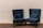 A pair of plush, dark blue velvet armchairs with high backs are positioned on a carpeted floor against a plain light beige wall. The chairs have decorative studded trim along the edges and are accompanied by matching lumbar pillows placed on the seat.