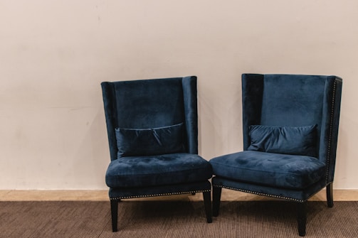 A pair of plush, dark blue velvet armchairs with high backs are positioned on a carpeted floor against a plain light beige wall. The chairs have decorative studded trim along the edges and are accompanied by matching lumbar pillows placed on the seat.