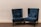A pair of plush, dark blue velvet armchairs with high backs are positioned on a carpeted floor against a plain light beige wall. The chairs have decorative studded trim along the edges and are accompanied by matching lumbar pillows placed on the seat.