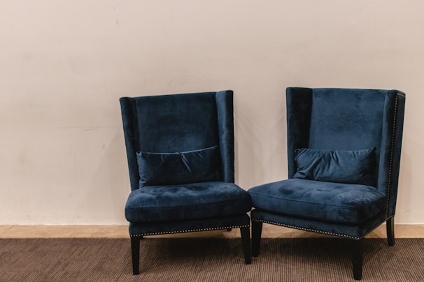 A pair of plush, dark blue velvet armchairs with high backs are positioned on a carpeted floor against a plain light beige wall. The chairs have decorative studded trim along the edges and are accompanied by matching lumbar pillows placed on the seat.