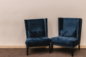 A pair of plush, dark blue velvet armchairs with high backs are positioned on a carpeted floor against a plain light beige wall. The chairs have decorative studded trim along the edges and are accompanied by matching lumbar pillows placed on the seat.