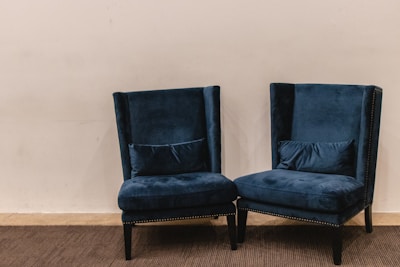 A pair of plush, dark blue velvet armchairs with high backs are positioned on a carpeted floor against a plain light beige wall. The chairs have decorative studded trim along the edges and are accompanied by matching lumbar pillows placed on the seat.