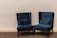 A pair of plush, dark blue velvet armchairs with high backs are positioned on a carpeted floor against a plain light beige wall. The chairs have decorative studded trim along the edges and are accompanied by matching lumbar pillows placed on the seat.