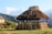 A rustic, thatched hut stands in a grassy area, with traditional wooden fencing around it. In the background, majestic snow-capped mountains rise against a clear blue sky. Two people are near the hut, admiring the view. Vibrant greenery and lush trees surround the scene.